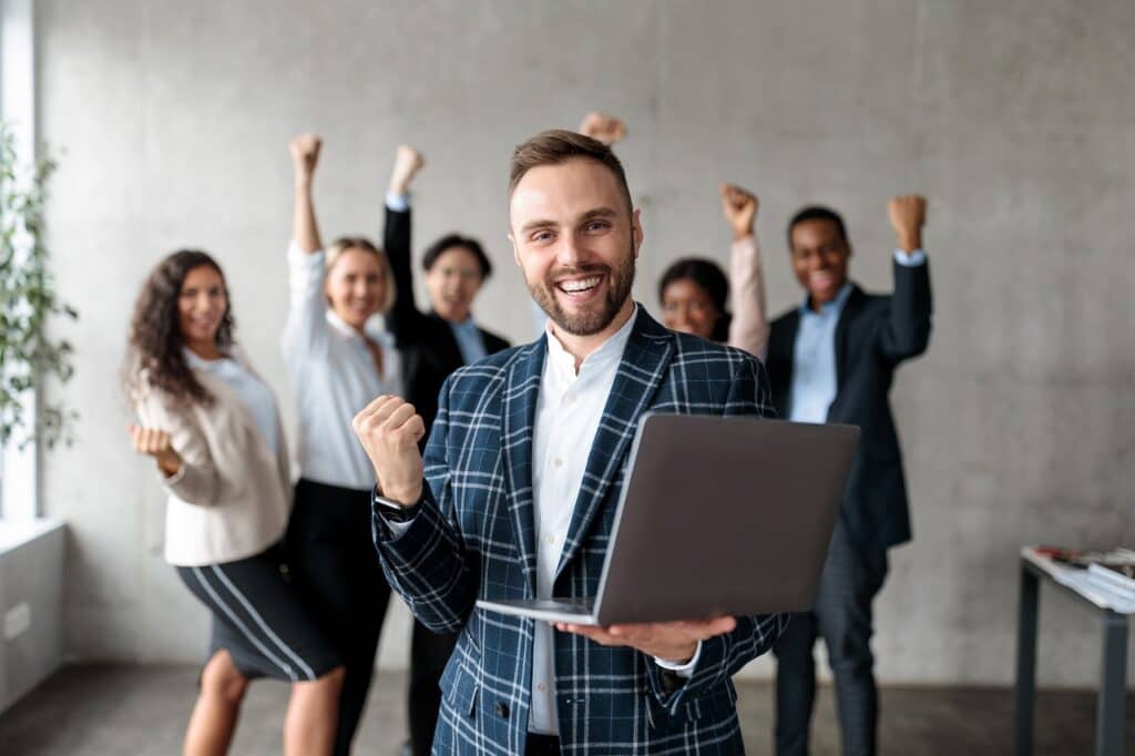 coding bootcamp Joyful Businessman With Laptop And His Business Team Celebrating Success At Work, Gesturing Yes Standing In Modern Office. Successful Internet Business And Startup, Joy Of Victory Concept