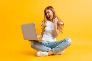 Young happy woman in white t-shirt is sitting using laptop and celebrating victory and success over yellow background