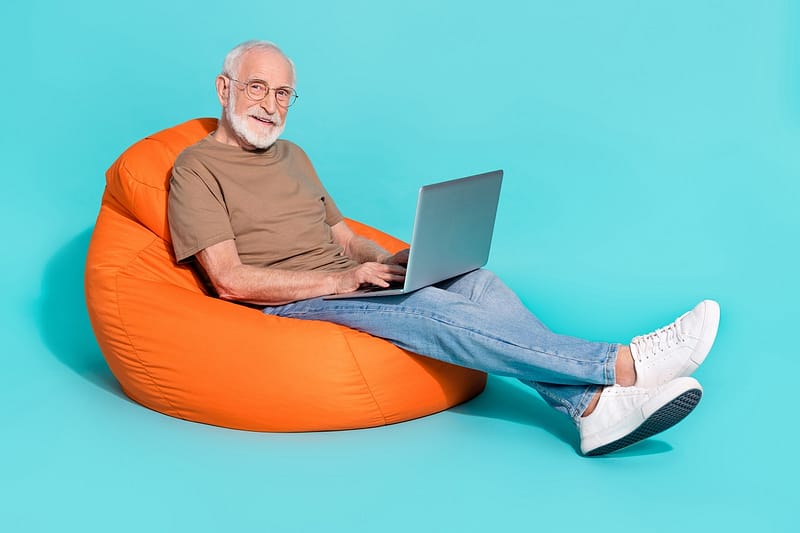 competitive as developer Portrait of handsome trendy cheery skilled grey-haired man sitting in bag using laptop isolated over bright blue color background.