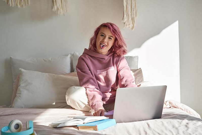 web development Cool hipster teen girl school gen z college student with pink hair wears hoodie sits on bed with laptop and book learning exam showing tongue with piercing looking at camera at home. Portrait.