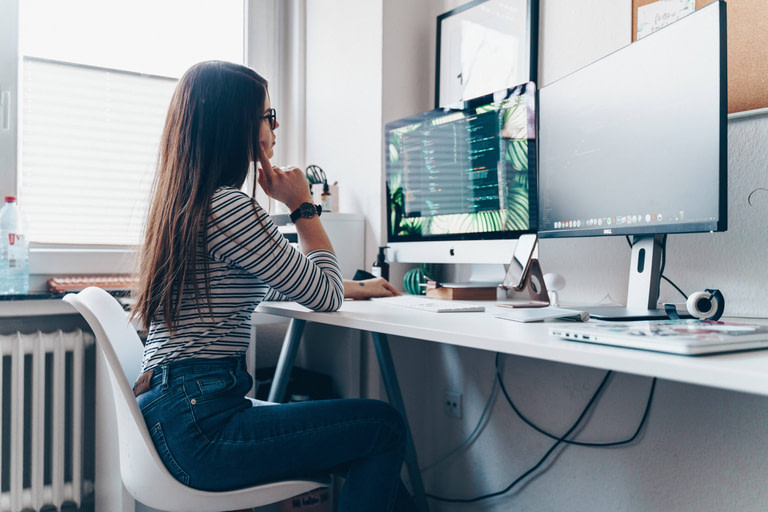 Young woman sitting at her desk in front of two screens learning how to code to become a web developer.